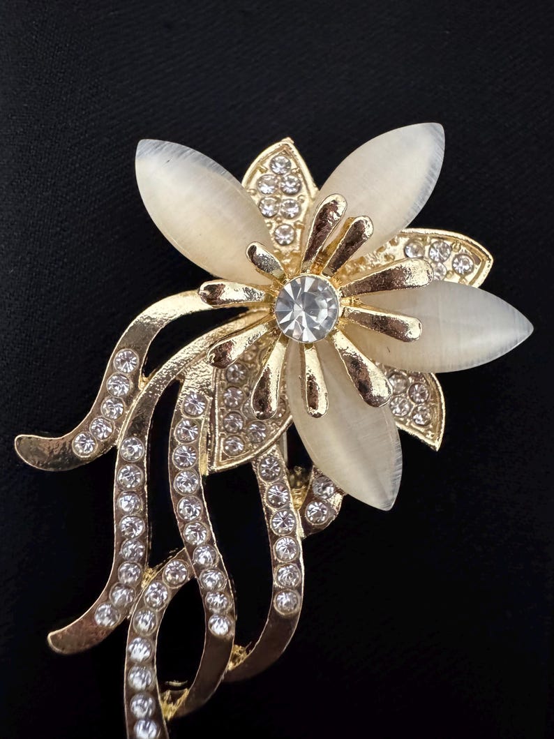 May include: A gold-toned flower brooch with a central clear crystal and cream-colored petals. The brooch features multiple clear crystals and curved gold accents, set against a black background.