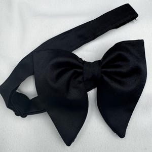 May include: A black bow tie featuring a large, symmetrical bow and an adjustable neckband. The fabric has a smooth, matte finish. The bow tie is displayed on a white surface, suitable for formal events.