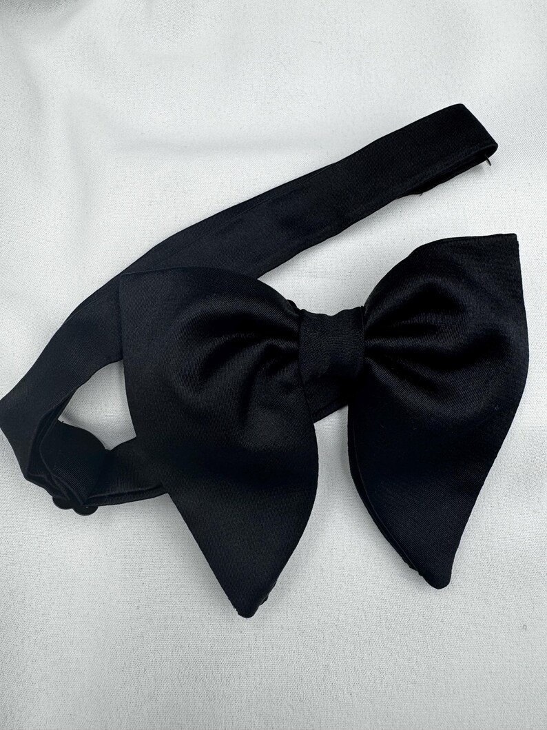 May include: A black bow tie featuring a large, symmetrical bow and an adjustable neckband. The fabric has a smooth, matte finish. The bow tie is displayed on a white surface, suitable for formal events.