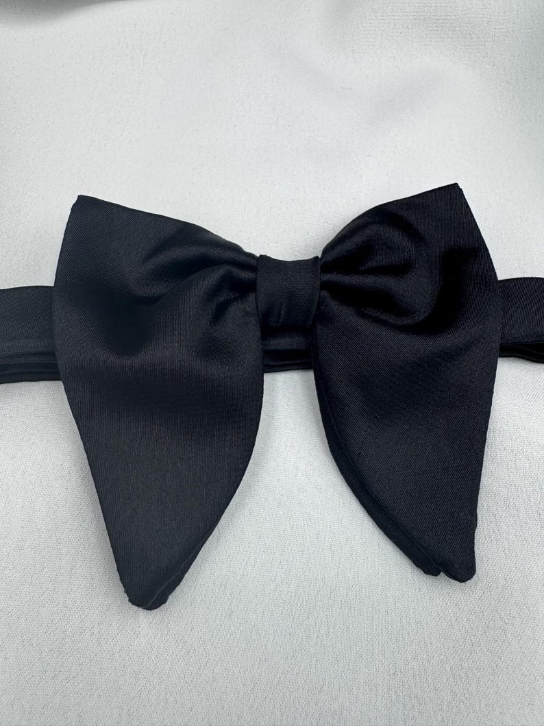 May include: A black bow tie, crafted from a smooth fabric, possibly satin. The image shows the bow tie from above, centered in the frame. This classic accessory is often worn with formal wear, such as a tuxedo or suit.