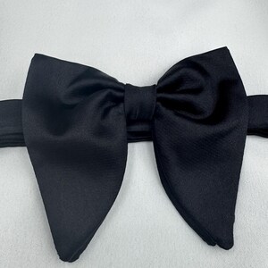 May include: A black bow tie made of satin, set against a white backdrop. The bow tie is centrally positioned, showcasing its symmetrical design and smooth, glossy texture. The material appears to be of a high quality.