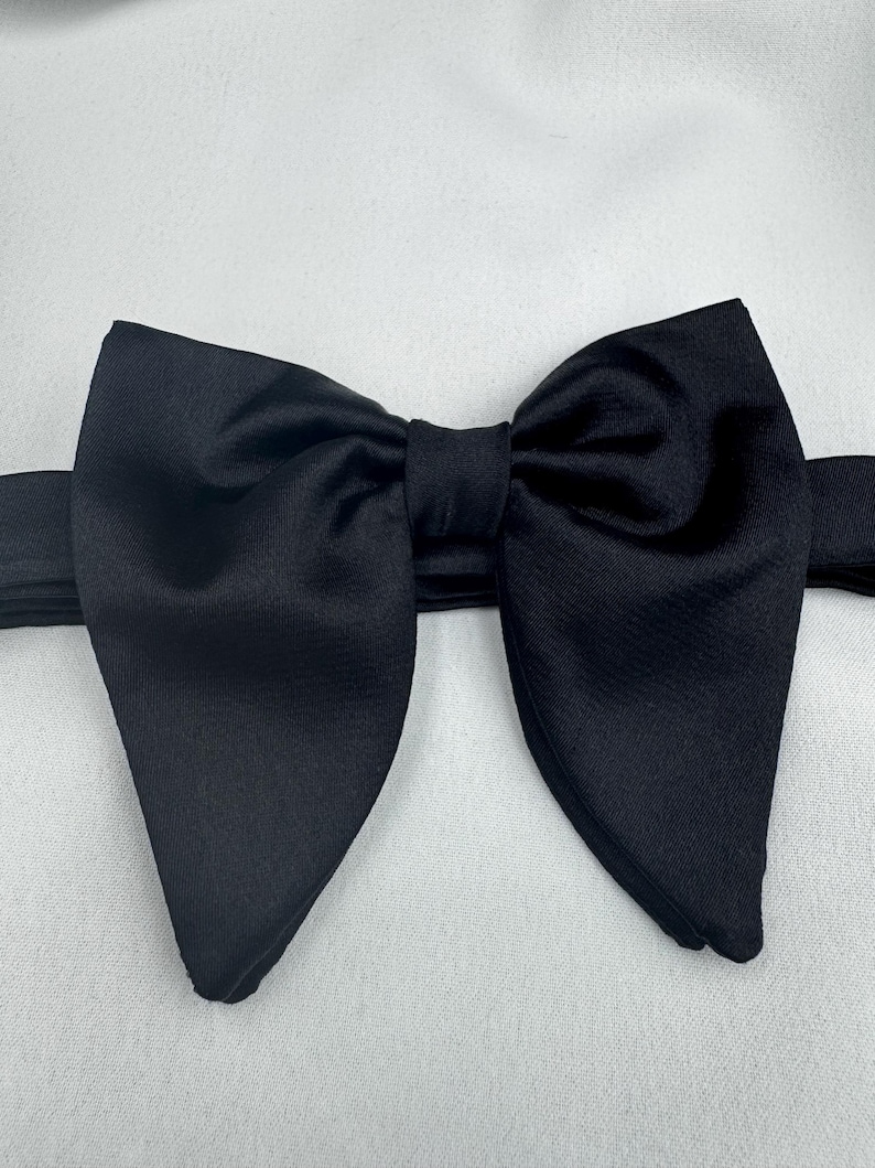 May include: A black bow tie made of satin, set against a white backdrop. The bow tie is centrally positioned, showcasing its symmetrical design and smooth, glossy texture. The material appears to be of a high quality.