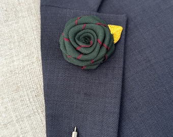 May include: A dark green fabric rose lapel pin with red accents and a small yellow leaf, attached to a navy blue blazer. A silver pin is also visible. This accessory adds a touch of elegance.