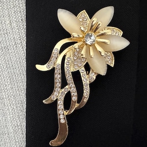 May include: A gold-toned floral brooch with a central clear rhinestone and cream-colored petals. The brooch features a flowing design with embedded clear rhinestones, set against a black background.