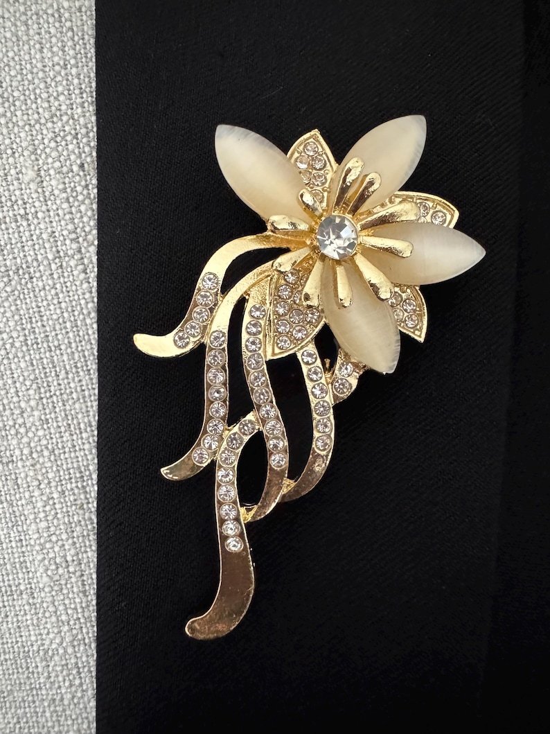 May include: A gold-toned floral brooch with a central clear rhinestone and cream-colored petals. The brooch features a flowing design with embedded clear rhinestones, set against a black background.
