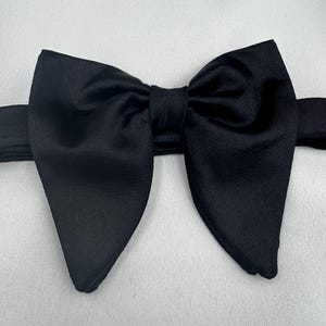 May include: A black bow tie, viewed closely. The bow tie is made from a smooth, glossy material and is tied in a traditional bow shape. It's a formal accessory, often worn with a tuxedo or suit for special occasions.