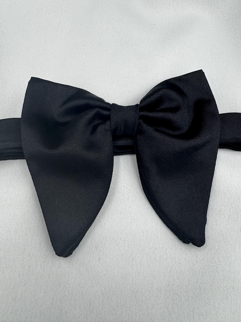 May include: A black bow tie, viewed closely. The bow tie is made from a smooth, glossy material and is tied in a traditional bow shape. It's a formal accessory, often worn with a tuxedo or suit for special occasions.