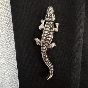 May include: A silver-colored alligator brooch pinned to a black fabric. The brooch is detailed with textured scales and small feet. The alligator is positioned diagonally, adding a touch of whimsy to the formal attire.