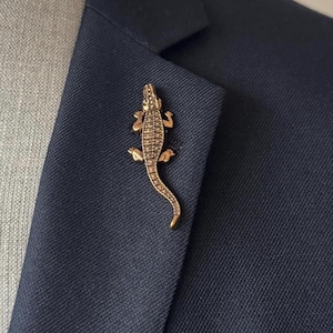 May include: A gold-colored alligator lapel pin on a navy blue blazer. The pin is detailed with textured scales and legs, and is attached to the lapel of the jacket. The alligator pin adds a touch of style to the blazer.