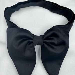 May include: A black satin bow tie with a matching elasticated band. The bow tie has a classic shape with pointed ends. The elasticated band is designed to fit around the neck. Suitable for formal events.