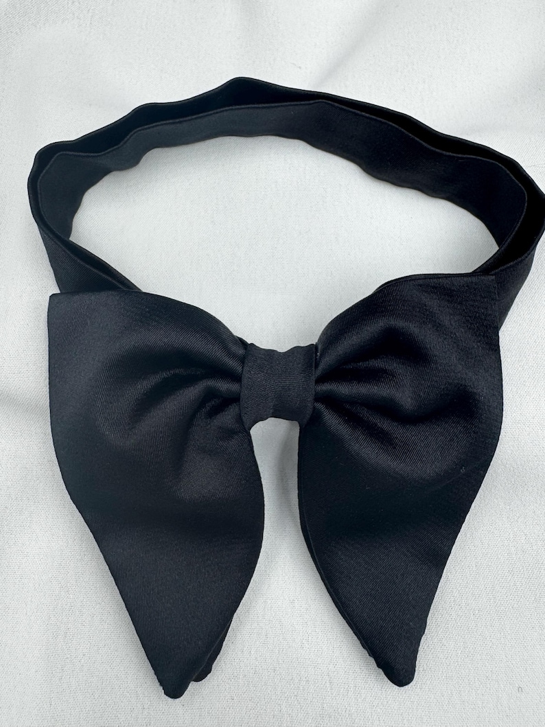 May include: A black satin bow tie with a matching elasticated band. The bow tie has a classic shape with pointed ends. The elasticated band is designed to fit around the neck. Suitable for formal events.