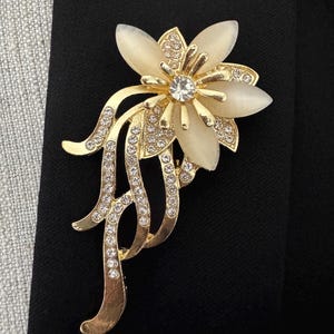May include: A gold-toned floral brooch with a central clear crystal and cream-colored petals. The brooch features delicate gold tendrils adorned with small, sparkling crystals. The brooch is set against a black background.