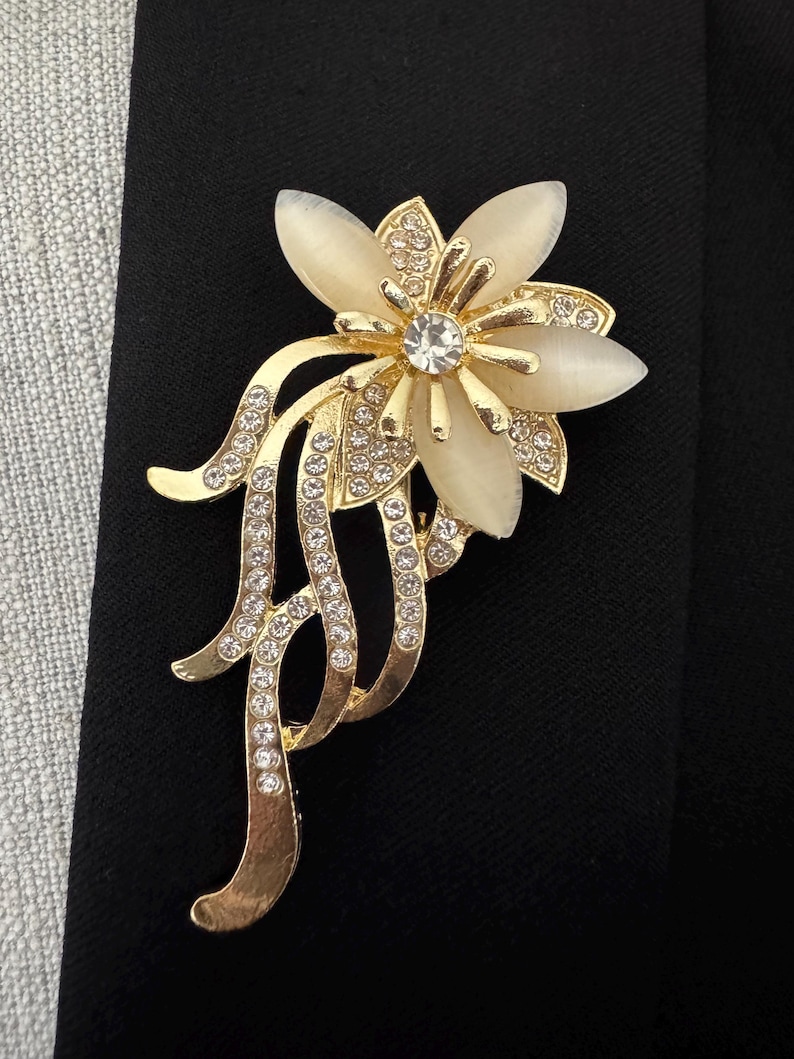 May include: A gold-toned floral brooch with a central clear crystal and cream-colored petals. The brooch features delicate gold tendrils adorned with small, sparkling crystals. The brooch is set against a black background.