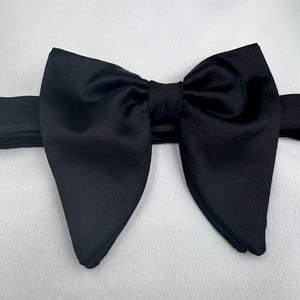 May include: A black bow tie, centrally positioned against a white backdrop. The bow tie is crafted from a smooth fabric, possibly satin. The bow is pre-tied, featuring a classic shape, with a band extending from each side.