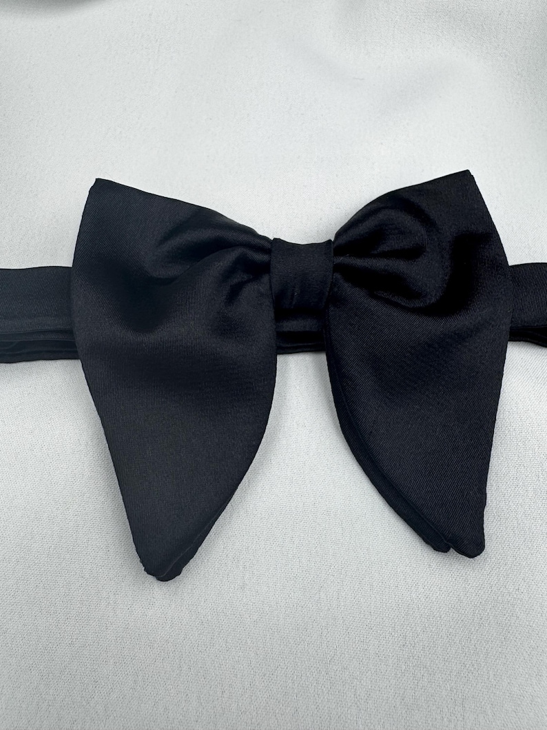May include: A black bow tie, centrally positioned against a white backdrop. The bow tie is crafted from a smooth fabric, possibly satin. The bow is pre-tied, featuring a classic shape, with a band extending from each side.