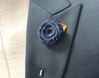 May include: A navy blue fabric rose lapel pin with a gold accent. The pin is worn on a dark blue suit jacket.