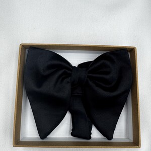 May include: A black bow tie made from a satin-like fabric, displayed in a rectangular cardboard box. The bow tie features a traditional design with a central knot and broad, symmetrical wings. The box has a white interior and a brown exterior.