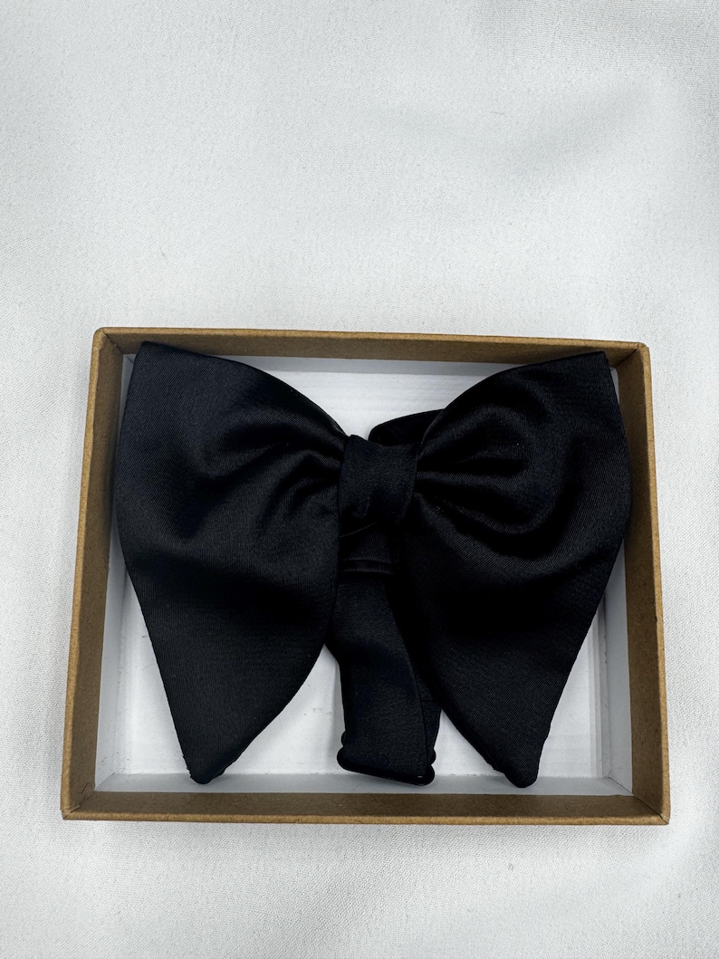May include: A black bow tie made from a satin-like fabric, displayed in a rectangular cardboard box. The bow tie features a traditional design with a central knot and broad, symmetrical wings. The box has a white interior and a brown exterior.