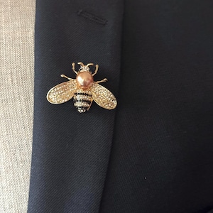 Puede incluir: Broche de abeja dorada con detalles de cristal en las alas y cuerpo perlado. La abeja está prendida a una blazer azul marino oscuro, creando un contraste llamativo. El broche añade un toque de elegancia.