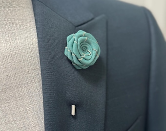 May include: A teal fabric lapel flower pin featuring a spiral rose design with subtle silver-toned thread accents. The floral accessory is attached to a dark navy blue suit jacket with a silver metal pin stem visible below the fabric flower.