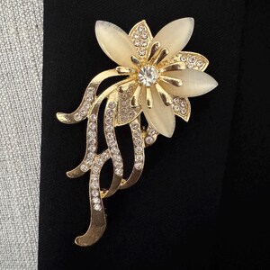 May include: A gold-toned floral brooch with a central clear crystal and cream-colored petals. The brooch features intricate detailing with small, sparkling crystals and flowing, curved accents. This elegant accessory is suitable for formal wear.