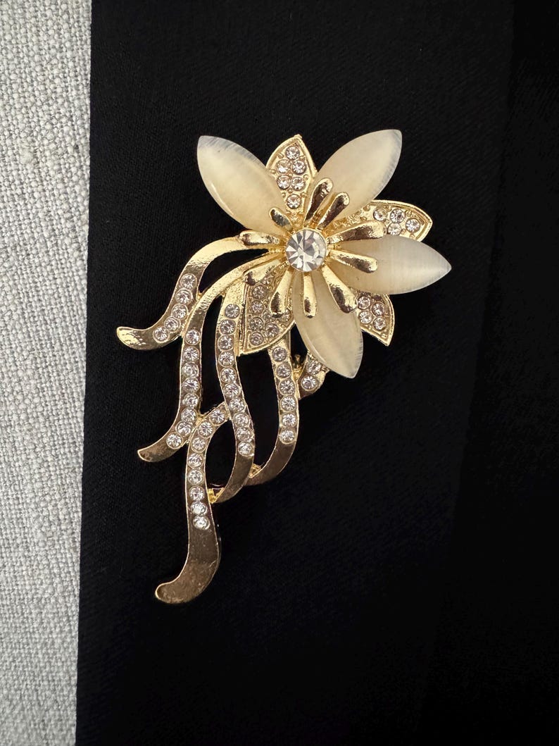 May include: A gold-toned floral brooch with a central clear crystal and cream-colored petals. The brooch features intricate detailing with small, sparkling crystals and flowing, curved accents. This elegant accessory is suitable for formal wear.