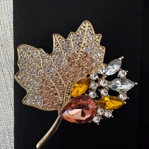 May include: A gold-toned maple leaf brooch featuring a textured surface encrusted with clear, champagne, and amber-colored rhinestones. The design includes a large pear-shaped peach crystal and several smaller marquise-cut amber and clear crystals.