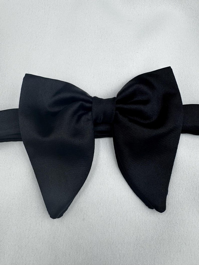 May include: A black bow tie, with a traditional butterfly design. The bow tie is crafted from a smooth, solid black material. The background is a plain white surface.