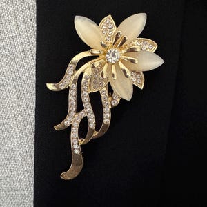 May include: A gold-toned floral brooch with a central clear rhinestone and cream-colored petals. The brooch features smaller clear rhinestones and a flowing, decorative design. The brooch is set against a black background.