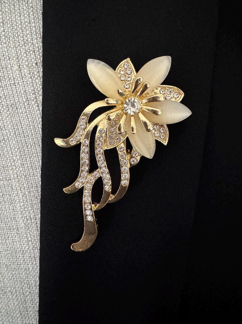 May include: A gold-toned floral brooch with a central clear rhinestone and cream-colored petals. The brooch features smaller clear rhinestones and a flowing, decorative design. The brooch is set against a black background.