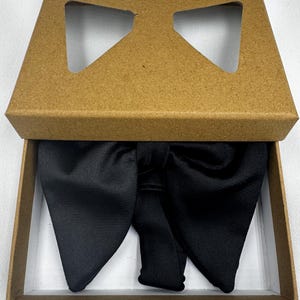 May include: A black bow tie presented in a brown cardboard box with a bow-shaped aperture in the lid. The bow tie is crafted from a smooth, lustrous material and is neatly arranged within the box.