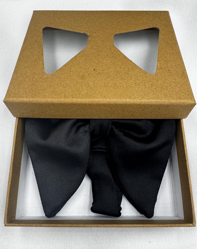 May include: A black bow tie presented in a brown cardboard box with a bow-shaped aperture in the lid. The bow tie is crafted from a smooth, lustrous material and is neatly arranged within the box.