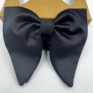 May include: A black satin bow tie, with a classic butterfly shape. The bow tie is neatly tied and appears to be new. It is presented on a brown cardboard box. The background is a plain white surface, providing a clean contrast.