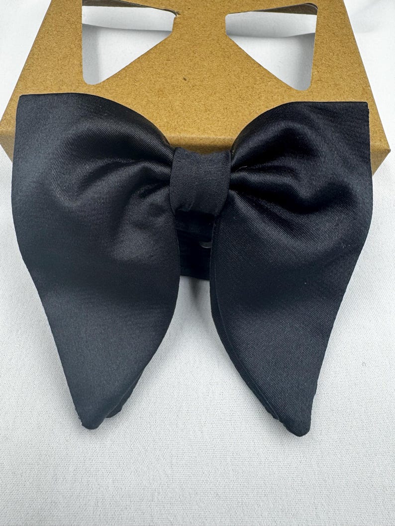 May include: A black satin bow tie, with a classic butterfly shape. The bow tie is neatly tied and appears to be new. It is presented on a brown cardboard box. The background is a plain white surface, providing a clean contrast.