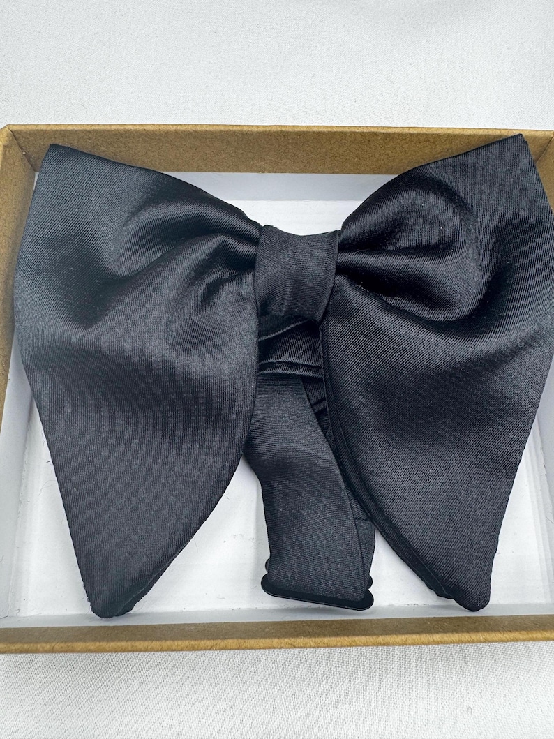 May include: A black bow tie made from a shiny material, presented in a cardboard box. The bow tie is neatly folded and ready to wear. A classic accessory for formal events.