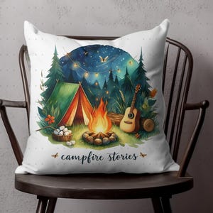 May include: A white throw pillow with a watercolor illustration of a campsite scene. The illustration features a tent, a campfire, a guitar, and a starry night sky. The text "campfire stories" is printed below the illustration.