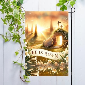 May include: A garden flag with a religious Easter theme. The flag features a stone tomb with an open doorway, two crosses, and a path leading away from the tomb. The flag is decorated with white flowers and the text "He is Risen".
