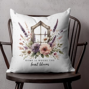 May include: White throw pillow with a watercolor floral design featuring lavender, pink, and purple flowers. A rustic wooden window frame is in the center of the pillow. The text "Home is where the heart blooms" is printed below the window.