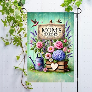 Könnte beinhalten: Eine Gartenflagge mit einem Blumendesign und einem Schild mit der Aufschrift "Mamas Garten". Die Flagge zeigt rosa und lila Blumen, eine Gießkanne und zwei Kolibris.