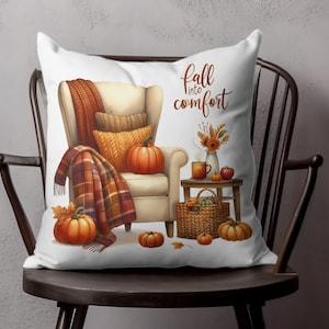 May include: A white throw pillow with a cozy autumn scene. An armchair with a blanket and pumpkins sits next to a small table with a mug and a basket of fruit. The text "fall into comfort" is printed on the pillow.