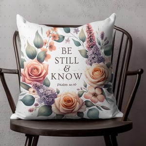 Op de afbeelding: Een wit kussen met een bloemenontwerp met rozen, lila en groen. Het kussen heeft de tekst "Be Still & Know" met de bijbelverwijzing "Psalm 46:10" erop gedrukt.
