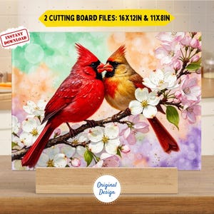 May include: A decorative cutting board with a painting of two cardinals on a flowering branch. The birds are red and yellow, with white blossoms and a soft, colourful background. The text "2 Cutting Board Files: 16x12in & 11x8in" is displayed. The dimensions are 40.6cm x 30.5cm and 27.9cm x 20.3cm.