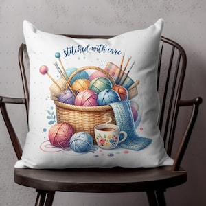 May include: White throw pillow with a watercolor illustration of a basket filled with yarn balls, knitting needles, and a blue knitted scarf. The pillow also features a floral teacup and the text "Stitched with care".