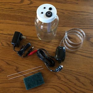 May include: A gold electroplating kit with a clear glass jar, a black power adapter, a clear plastic tube, two metal rods, a green scouring pad, and a black air pump.