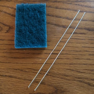 May include: Two silver metal skewers and a blue cleaning sponge on a wooden surface.