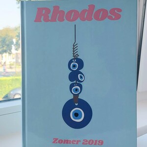 May include: A light blue book with the title "Rhodos" in pink lettering. The book features a blue and white evil eye charm hanging from a string. The text "Zomer 2019" is printed in pink at the bottom of the book.