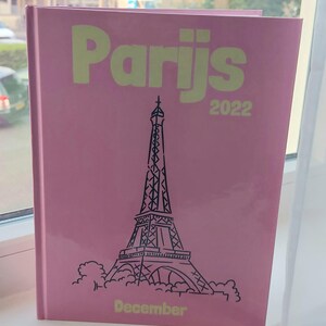 May include: A pink planner with the word "Parijs" in yellow letters and the year "2022" in black letters. The planner features a black and white illustration of the Eiffel Tower and the word "December" in yellow letters.