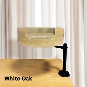 Puede incluir: Una cama para gatos montada en la pared de madera de roble blanco. La cama tiene un dise&ntilde;o curvo y abierto con un acabado de veta de madera clara. Est&aacute; soportada por un brazo y una abrazadera de metal negro, fijados a un escritorio de madera. El texto "White Oak" se muestra en la parte inferior.