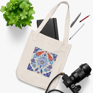 May include: Cream-colored tote bag featuring a square, colorful floral design with red tulips and blue accents. The bag has long handles and is made of a sturdy fabric. A black notebook, pens, and a camera are also visible.