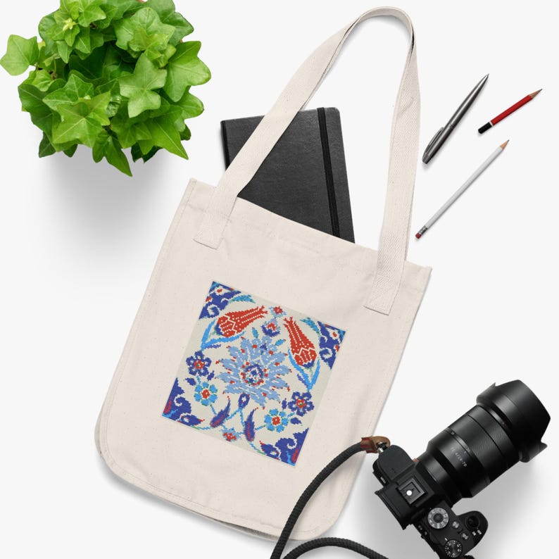 May include: Cream-colored tote bag featuring a square, colorful floral design with red tulips and blue accents. The bag has long handles and is made of a sturdy fabric. A black notebook, pens, and a camera are also visible.